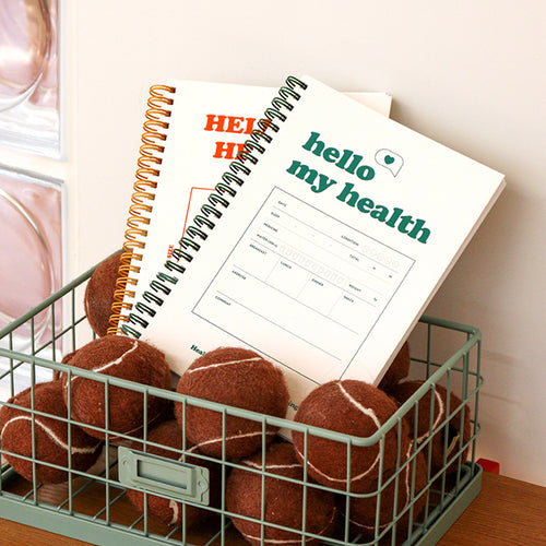 A metal wire basket filled with brown tennis balls holds two spiral-bound health journals. One journal has an orange “HELLO MY HEALTH” title, and the other has a green “hello my health” title with a health tracking form visible on the cover.
